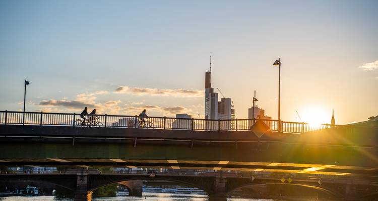Cyclists crossing a bridge in Frankfurt silhouetted against a warm sunset with the city skyline in the background