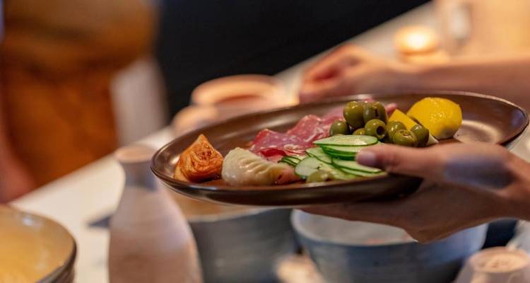 Nahaufnahme einer Hand, die einen Keramikteller mit verschiedenen Vorspeisen hält, darunter Oliven, Räucherfleisch und Gemüse.