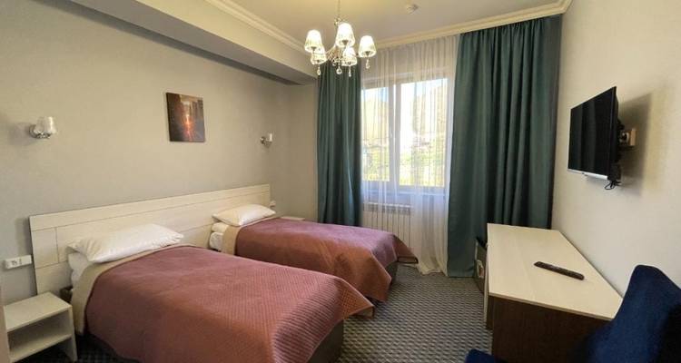 Einfaches Hotelzimmer mit zwei Einzelbetten, neutraler Einrichtung, Kronleuchterbeleuchtung und einem Fenster mit blaugrünen Vorhängen.