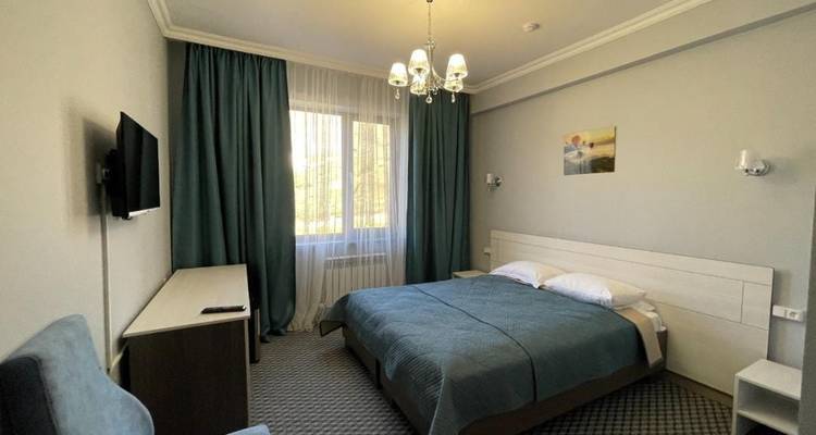Komfortables Doppelhotelzimmer mit Kingsize-Bett, an der Wand montiertem Fernseher und großem Fenster mit grünen Vorhängen.