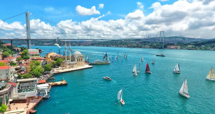Lebendige Luftaufnahme der Bosporus-Straße in Istanbul mit der Ortaköy-Moschee, Segelbooten und Hängebrücke an einem sonnigen Tag.