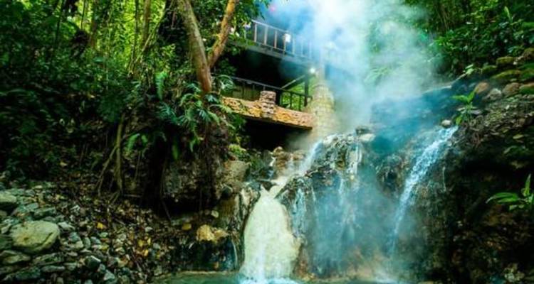 El vapor sube de una pequeña cascada tropical y una piscina natural debajo de un puente de madera en la exuberante selva.