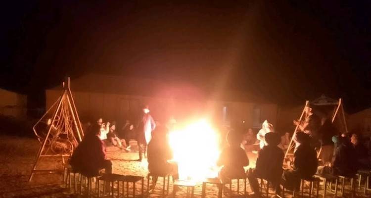 Les gens s'assoient sur de petits tabourets autour d'un grand feu de camp dans un camp du désert sous le ciel nocturne.