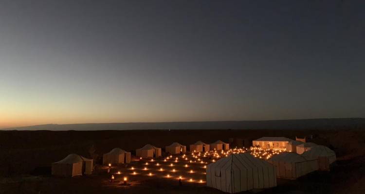 Panorama nocturne de tentes blanches du désert illuminées par de nombreuses lanternes au sol sous un ciel crépusculaire.