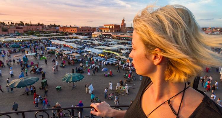 Voyageuse blonde regardant la foule animée du soir de la place Jemaa el-Fnaa depuis une terrasse sur le toit.