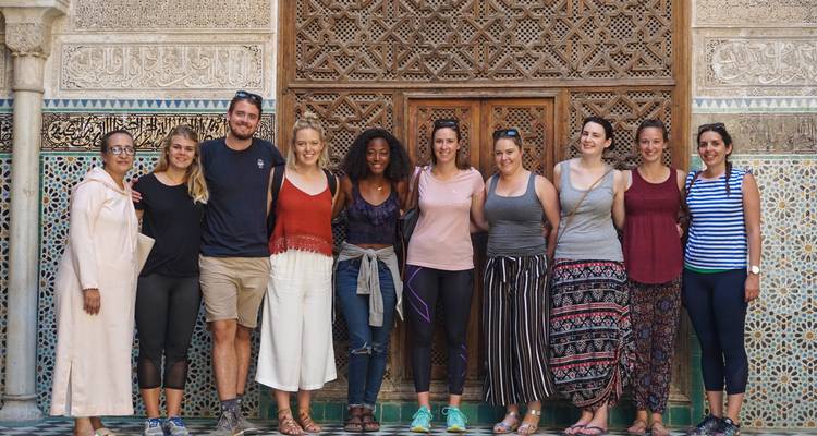Groupe de touristes posant devant les carrelages complexes et la porte en bois sculpté d'une médersa historique.