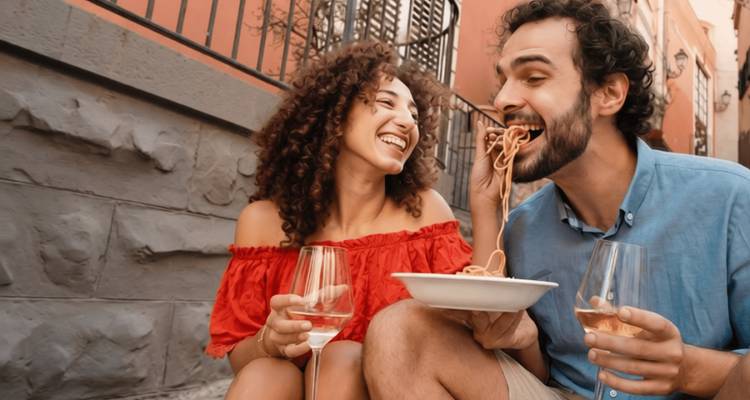 Laughing couple sharing spaghetti and white wine outside a colorful Italian alley.