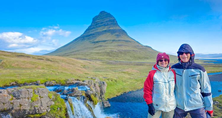 Couple in jackets posing beside a small waterfall with Kirkjufell mountain rising behind them under a blue sky.