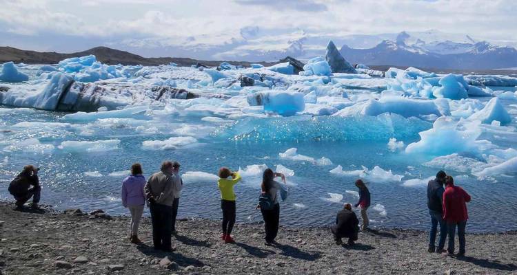 Travelers exploring the shore of a vivid blue glacial lagoon dotted with icebergs and distant snowy peaks.