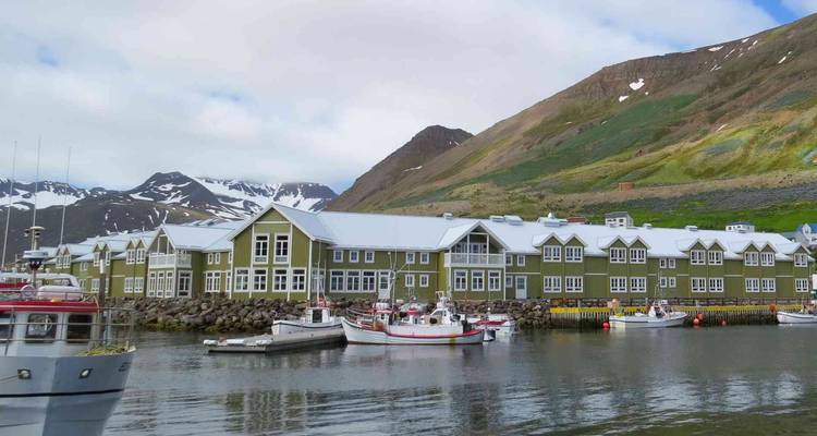 Quaint harbor town with green wooden buildings, boats moored on calm water, and snow-tipped hills behind.