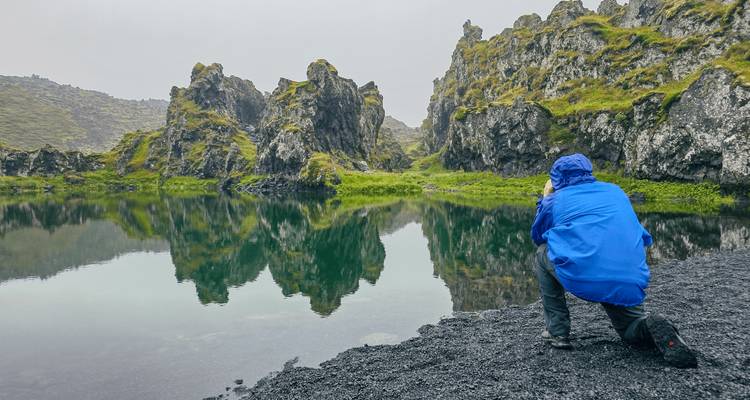 Photographer in blue jacket kneeling on a black-sand shore capturing reflections of jagged lava rocks in still water.