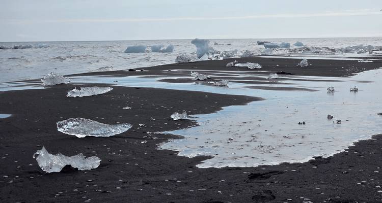 Black-sand beach scattered with clear ice chunks beside a calm grey sea under soft light.