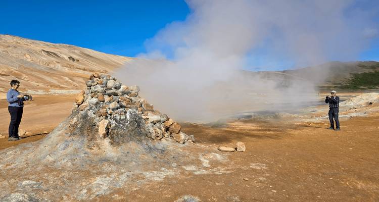 Steam billows from a rocky geothermal vent in a barren ochre landscape while two visitors take photos.