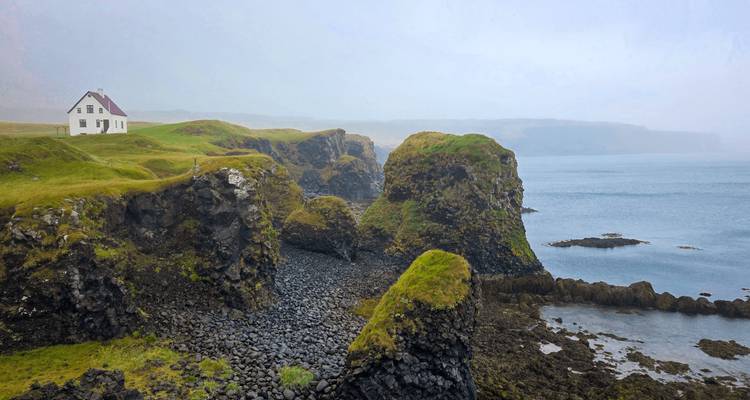 Dramatic sea cliffs with a solitary white house perched on a grassy promontory above calm blue water.
