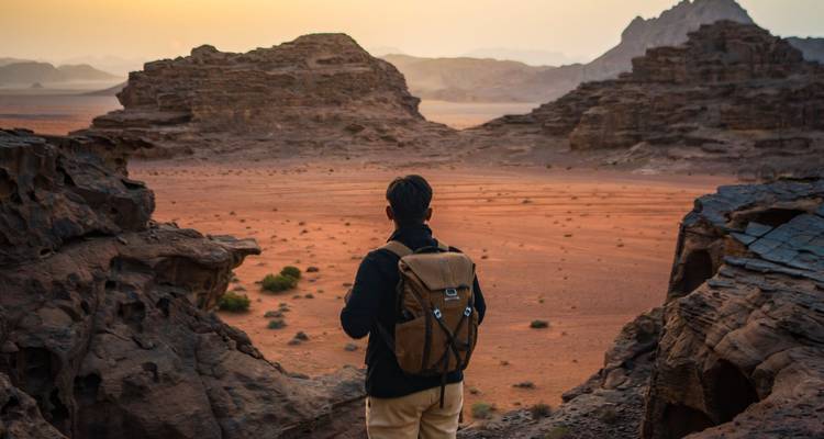 Un mochilero viajando solo contempla las formaciones de arenisca esculpidas por el viento y los macizos rocosos distantes de Wadi Rum al amanecer.