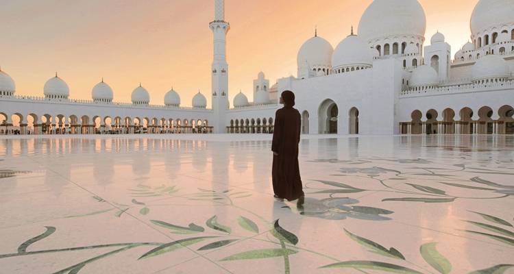 Eine einzelne Gestalt spaziert bei Sonnenuntergang über den glänzenden Marmor des Innenhofs der Sheikh Zayed Großmoschee.