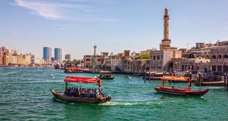 Traditionelle hölzerne Abras, die den Dubai Creek überqueren, mit historischen Gebäuden und moderner Skyline im Hintergrund.