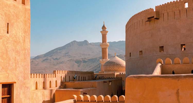 Blick über die Wehrgänge der Nizwa-Festung auf eine Moschee mit Kuppel und eine entfernte Bergkette unter klarem Himmel.