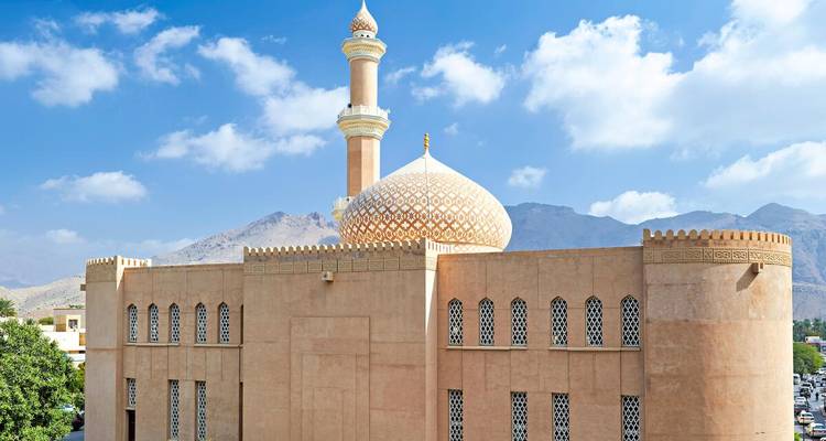 Moschee in Beige mit gemusterten Fenstern und verzierter Kuppel vor rauen Bergen und blauem Himmel.