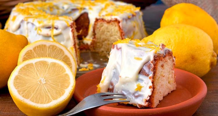 Tranche de gâteau au citron moelleux avec glaçage crémeux entourée de citrons frais sur une table rustique.
