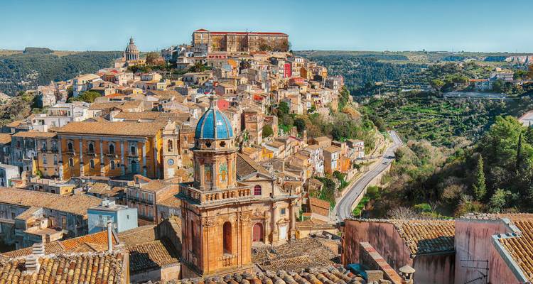 Vue panoramique sur les bâtiments baroques colorés de Ragusa Ibla et son dôme aux tuiles bleues, entourés de collines.