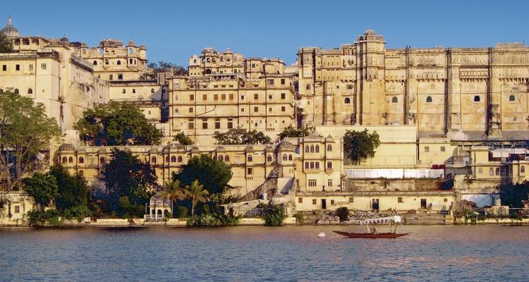 Seefassade des Udaipur City Palace, die im Morgenlicht erstrahlt, mit einem kleinen Boot auf dem Wasser.