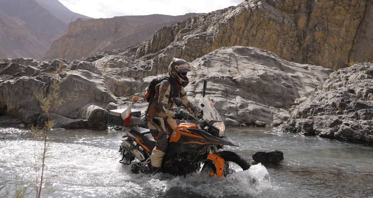 Ein Motorradfahrer in vollständiger Schutzausrüstung fährt durch einen flachen Bergbach, umgeben von rauen Felsklippen in Pakistan.