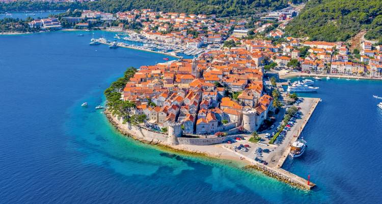 Vista aérea de un pueblo medieval amurallado con edificios de techos naranjas en una pequeña isla del Adriático rodeada de agua turquesa y un puerto deportivo