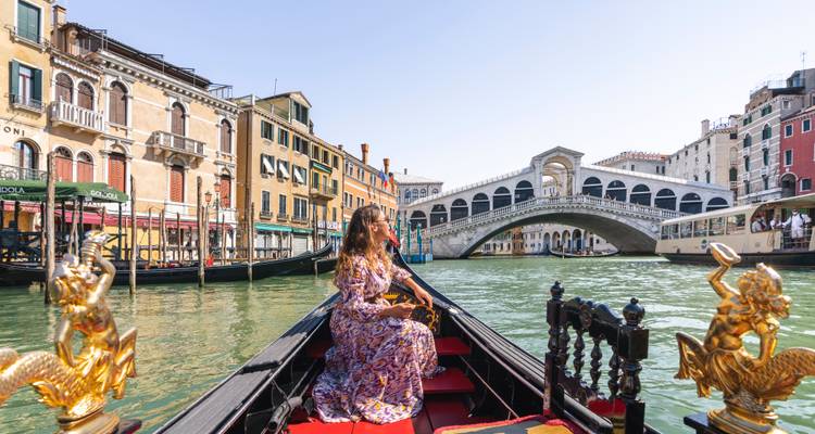 Une femme en robe à fleurs navigue sur une gondole décorée avec le Pont du Rialto se dressant majestueusement à l'arrière-plan sur le Grand Canal