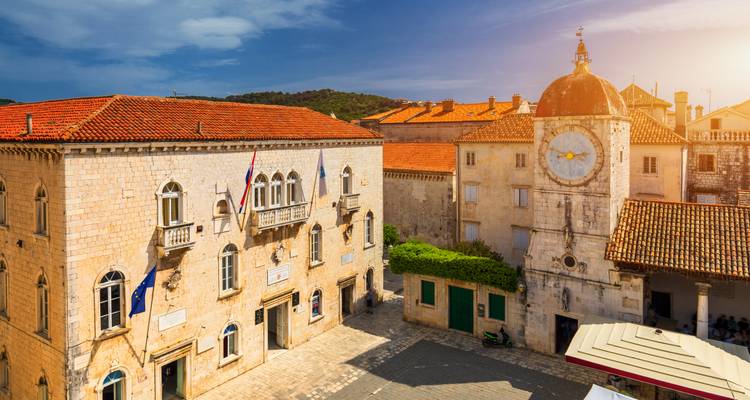 Plaza soleada en Trogir con torre de reloj de piedra y edificios históricos.
