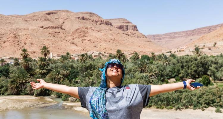 Le voyageur étend les bras largement devant une vallée luxuriante remplie de palmiers encadrée par des montagnes marocaines arides.