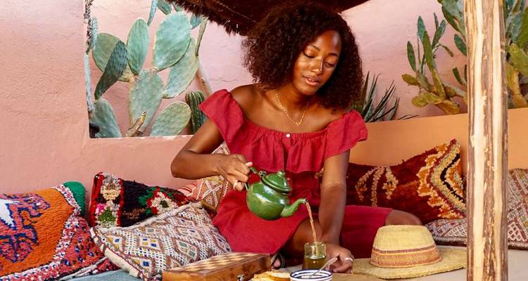 Une femme dans un riad coloré verse un thé à la menthe fumant dans un petit verre parmi des coussins patterned et des cactus.