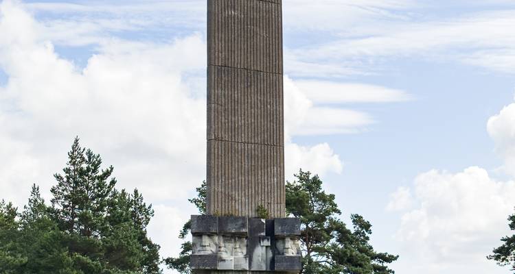 Colonne commémorative en béton imposante s'élevant au-dessus des cimes des arbres, avec des visages sculptés à sa base sur un fond de ciel bleu.