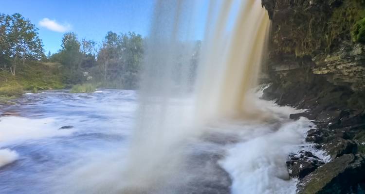 Puissante cascade observée de derrière un rideau d'eau ambrée et tourbillonnante versant dans une rivière mousseuse en contrebas.