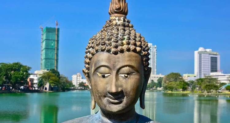 Estatua de Buda de bronce mirando hacia un lago tranquilo con los edificios de la ciudad de Colombo de fondo.