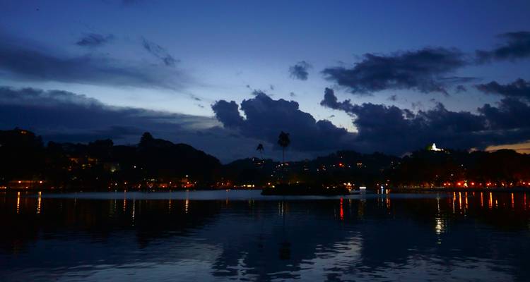 Vista al atardecer del Lago Kandy con colinas recortadas contra el cielo y reflejos de las luces de la ciudad.