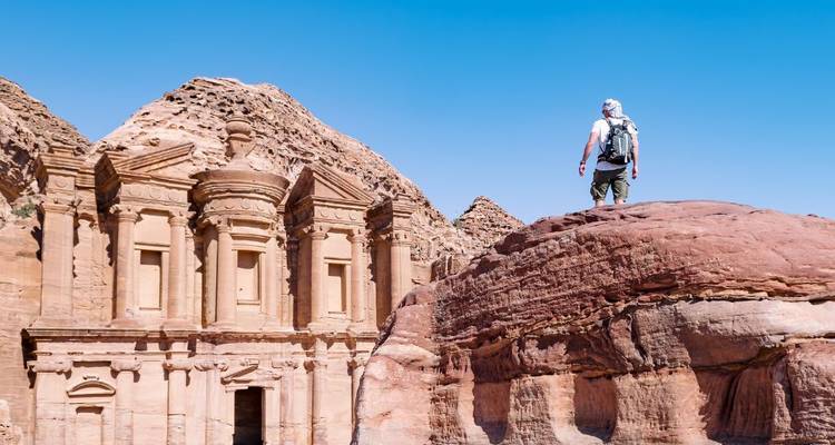 Backpacker standing on a red sandstone ridge facing the massive façade of Petra’s Monastery under a clear blue sky.