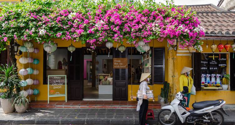 Een met bloemen versierde gele winkelvoorgevel in Hoi An met lantaarns die eroven hangen en een lokale vrouw in een puntige hoed die naast een scooter staat.