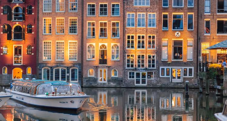 Les lumières du soir brillent sur les maisons en briques bordant les canaux tandis qu'un bateau touristique glisse dans le centre d'Amsterdam.