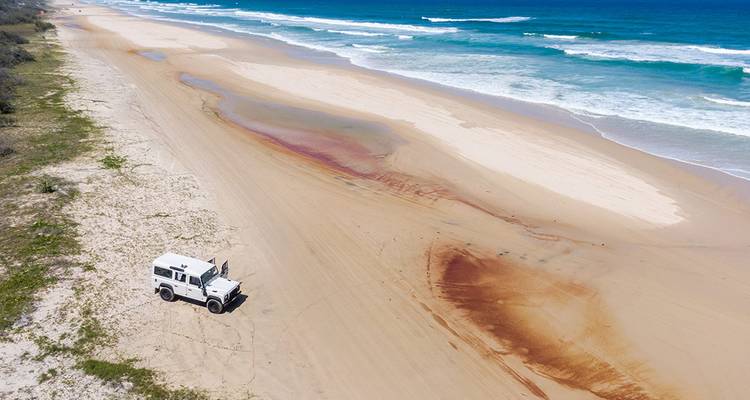 Un véhicule 4x4 garé sur une vaste plage dorée à côté des vagues de l'océan qui roulent sous un ciel lumineux.
