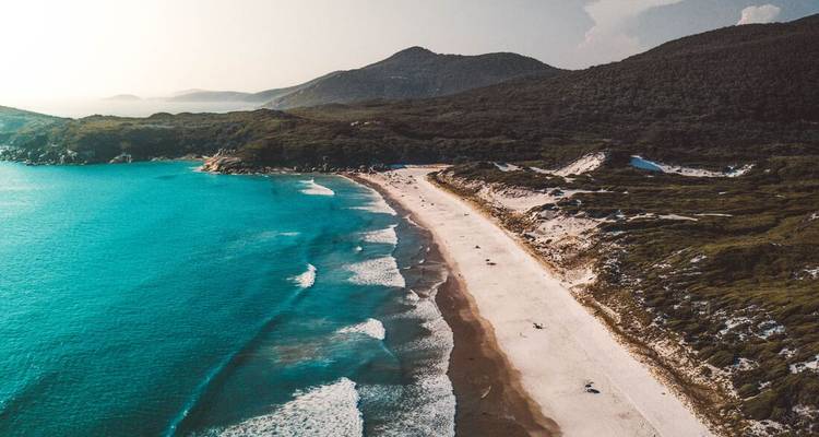 Dronebeeld van een turquoise baai en een weelderig wit zandstrand omgeven door rotsachtige heuvels in het licht van de late middag.