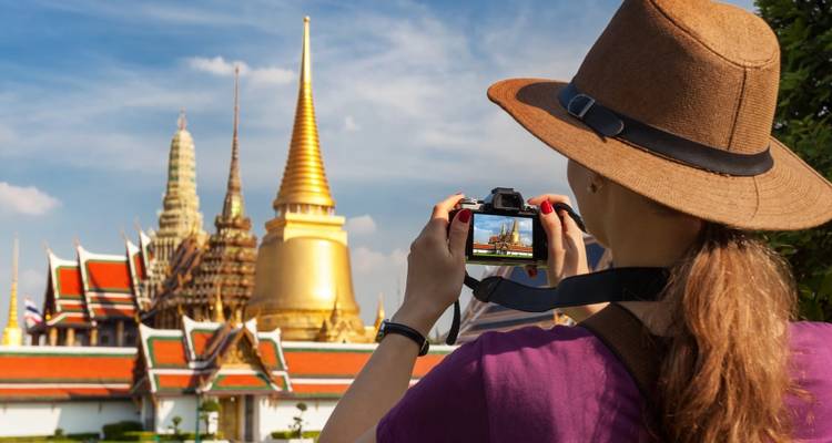 Mujer joven con sombrero fotografiando las doradas agujas del Gran Palacio de Bangkok contra un cielo azul