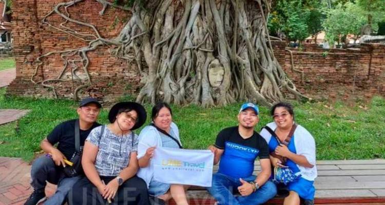 Grupo sonriente de turistas posando con un cartel frente a la cabeza de Buda incrustada en el árbol en Ayutthaya