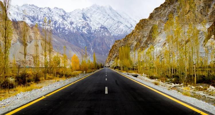 Straight black highway stretches toward snow-capped peaks, flanked by golden poplars and rocky cliffs.