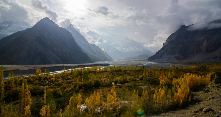 Autumn trees line a broad valley floor beneath layers of misty mountains and dramatic cloud cover.