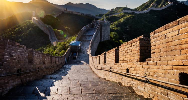 Sun-lit stone steps of the Great Wall of China winding over green mountains.