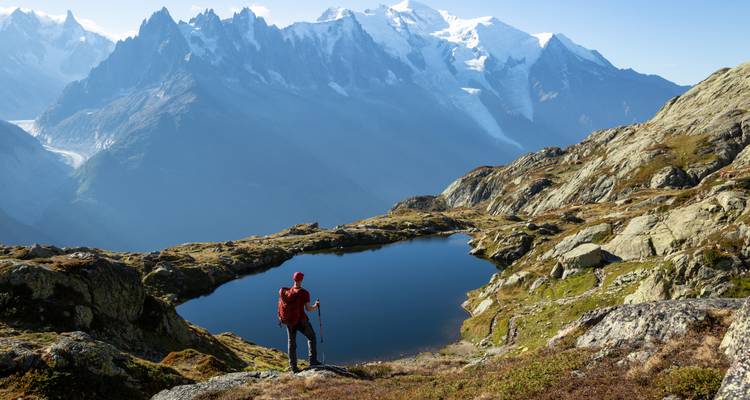 Un randonneur solitaire en veste rouge se tient à côté d'un lac alpin face aux pics enneigés de la chaîne du Mont-Blanc.