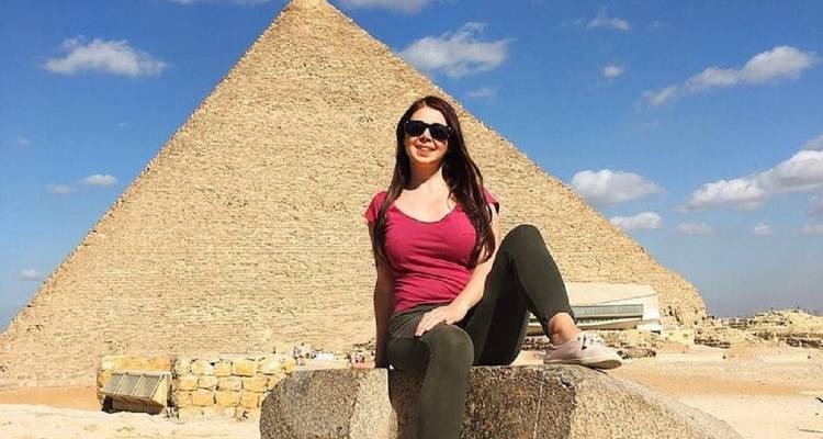 Femme assise sur un bloc de pierre souriante avec la Grande Pyramide s'élevant derrière elle sous un ciel bleu éclatant.