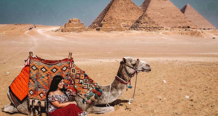 Voyageur vêtu d'une robe à motifs assis à côté d'un chameau orné avec les trois pyramides de Giza en arrière-plan sablonneux.