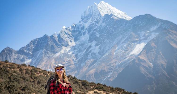 Una trekkista posa bajo un imponente pico nevado del Himalaya bajo un cielo azul despejado.